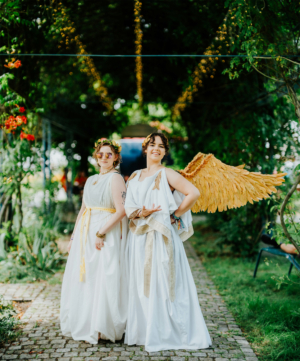 Zwei Frauen in eleganten, weißen Kostümen stehen auf einem gepflasterten Weg unter grünen Bäumen. Eine trägt goldene Flügel, während die andere eine Sonnenbrille und Schmuck trägt. Die Atmosphäre ist festlich und kreativ, passend zum Elbenwald Festival.