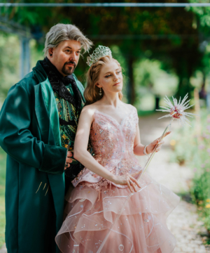 Ein Paar in fantasievollen Kostümen, inspiriert von Märchen, posiert im Freien. Die Frau trägt ein glitzerndes, rosa Kleid mit Rüschen und eine Krone, während der Mann einen eleganten, grünen Umhang trägt. Die Szene ist von bunten Blumen umgeben, die eine festliche Atmosphäre schaffen.