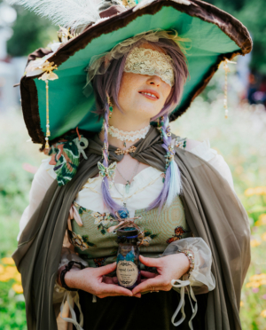 Frau in fantasievoller Kostümierung mit breitem Hut und verzierten Accessoires, die ein Fläschchen mit der Aufschrift „Liquid Luck“ hält. Im Hintergrund blühende Wiesen, die die kreative Atmosphäre des Elbenwald Festivals unterstreichen.