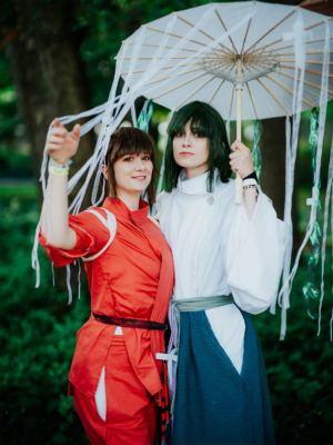 Zwei Cosplayerinnen in bunten Kostümen posieren fröhlich unter einem japanischen Schirm beim Elbenwald Festival. Die Szene zeigt die kreative Atmosphäre des Festivals, das Musik und Nerdkultur vereint, umgeben von grüner Natur.
