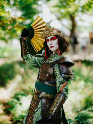 Eine cosplaying Person in detaillierter Rüstung mit rotem Make-up und einem Fächer, posiert im Freien auf dem Elbenwald Festival, umgeben von grüner Natur und sanften Lichtverhältnissen.