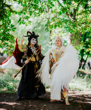 Zwei kostümierte Teilnehmer auf dem Elbenwald Festival stehen im Freien. Die erste Person trägt ein beeindruckendes schwarzes und rotes Kostüm mit Hörnern und einem Zepter, während die zweite Person in einem eleganten weißen Gewand mit großen Flügeln gekleidet ist.