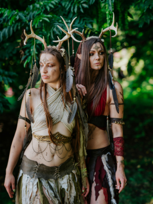 Zwei Frauen in fantasievollen Kostümen mit Geweih-Accessoires und Körperbemalung stehen im Grünen. Ihre Outfits sind kreativ gestaltet und spiegeln den mystischen Charakter des Elbenwald Festivals wider, das Musik und Nerdkultur vereint.