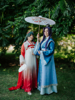 Zwei Cosplayerinnen in farbenfrohen, traditionellen Kostümen stehen unter einem Baum, während eine von ihnen einen Sonnenschirm hält. Die Szene fängt die kreative Atmosphäre des Elbenwald Festivals ein, das Musik und Geek-Kultur vereint.