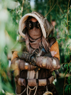 Ein kostümierter Besucher des Elbenwald Festivals steht zwischen grünen Pflanzen. Er trägt eine auffällige Maske, eine Kapuze und gepanzerte Accessoires, die seinen einzigartigen Look unterstreichen. Die Atmosphäre ist kreativ und fantasievoll.