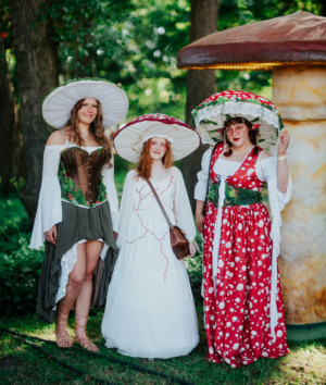 Drei Frauen in fantasievollen Pilzkostümen stehen im Freien, umgeben von Bäumen. Die Outfits sind kreativ gestaltet, mit großen, dekorativen Hüten und farbenfrohen Kleidern, die an die Natur und Märchen erinnern. Ideal für das Elbenwald Festival.