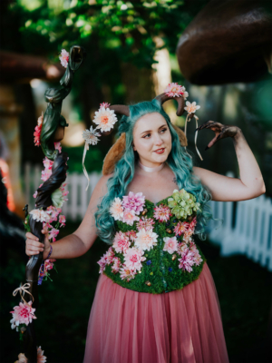 Eine Frau mit blauen Haaren und einem blumengeschmückten, grünen Oberteil posiert auf dem Elbenwald Festival. Sie hält einen dekorierten Stock und trägt einen pinken Rock, umgeben von einer grünen, natürlichen Kulisse mit Pilzen im Hintergrund.