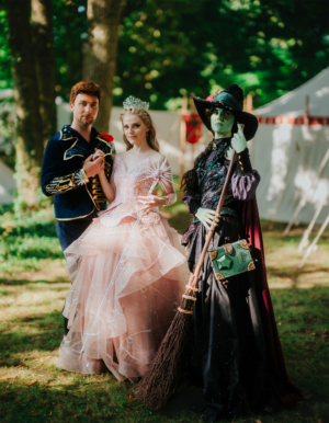 Drei verkleidete Teilnehmer des Elbenwald Festivals posieren in einem grünen Wald. Ein Prinz in blauer Uniform, eine Prinzessin in glitzerndem rosa Kleid und eine Hexe mit grüner Haut und Besen zeigen ihre fantasievollen Kostüme vor einem Zelt.