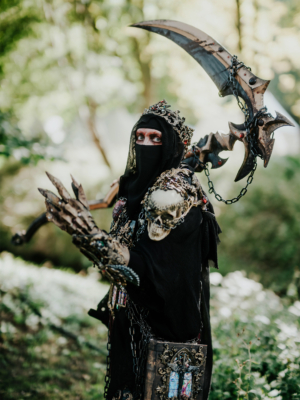 Ein kostümierter Teilnehmer des Elbenwald Festivals, verkleidet als mystische Figur mit einer beeindruckenden Waffe und detailreicher Rüstung, steht in einer grünen Umgebung. Der Fokus liegt auf dem kreativen Outfit und der fantasievollen Darstellung.