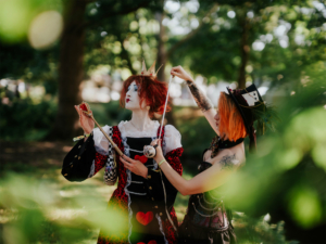 Zwei Cosplayerinnen in fantasievollen Outfits auf dem Elbenwald Festival, eine verkleidet als Herz-Königin mit rotem Kleid und Krone, die andere in einem schwarzen Korsett mit auffälligen Tattoos. Die Szene spielt sich in einer grünen, lebhaften Umgebung ab.