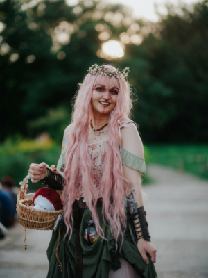 Eine kostümierte Teilnehmerin des Elbenwald Festivals mit langen, rosa Haaren und einem Blumenkranz lächelt in die Kamera. Sie hält einen Korb mit einem Plüschspielzeug und trägt ein fantasievolles Outfit, das von der Natur inspiriert ist. Hintergrund mit unscharfem Grün.