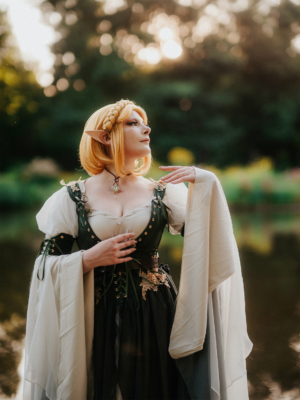 Eine cosplaying Frau in einem eleganten, elfischen Outfit mit blonden Haaren steht am Ufer eines Gewässers. Die Szenerie ist von sanftem Licht durchflutet, was eine magische Atmosphäre schafft, passend zur kreativen Stimmung des Elbenwald Festivals.