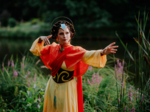 Eine cosplayende Person in einem farbenfrohen Kostüm steht vor einem Gewässer, umgeben von grünen Pflanzen und lila Blumen. Die Pose deutet auf eine magische oder kämpferische Handlung hin, passend zum kreativen Flair des Elbenwald Festivals.