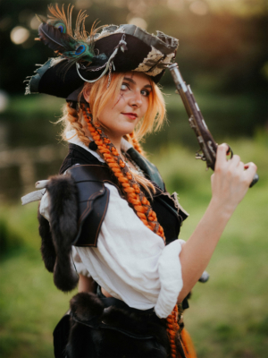 Cosplayerin mit orangefarbenen Zöpfen und Piratenkostüm, inklusive Federhut und Pistole, posiert vor einer grünen Kulisse beim Elbenwald Festival. Die Szene fängt die kreative Atmosphäre und den Spaß des Festivals ein.