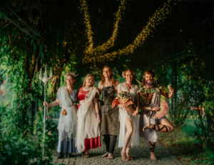 Fünf kreative Kostüme, inspiriert von Mythologie und Fantasie, auf dem Elbenwald Festival. Die Teilnehmer posieren unter einer mit Lichtern geschmückten Pergola, umgeben von üppigem Grün, und zeigen die lebendige Atmosphäre des Festivals.