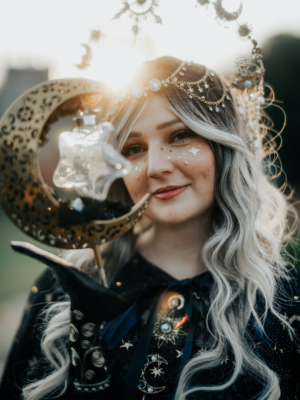 Eine kostümierte Person mit einer Sternen- und Monddekoration hält eine glitzernde Flasche in der Hand. Im Hintergrund strahlt die Sonne, während die Person mit funkelndem Make-up und einem eleganten Outfit im Elbenwald Festival posiert.