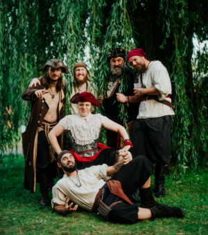 Sechs verkleidete Piraten posieren fröhlich vor einem Weidenbaum beim Elbenwald Festival, einem Mix aus Musikfestival und Convention, und zeigen ihre kreativen Kostüme und den gemeinschaftlichen Spaß.