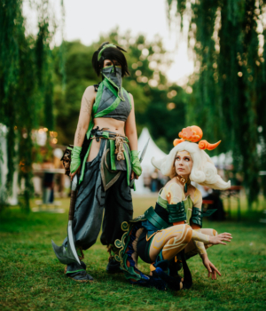 Zwei Cosplayer in beeindruckenden Kostümen auf dem Elbenwald Festival. Die erste Person trägt ein grünes und graues Outfit mit Waffen, während die zweite in einem bunten, detailreichen Kostüm mit charakteristischem Haar posiert. Hintergrund mit Festivalatmosphäre.