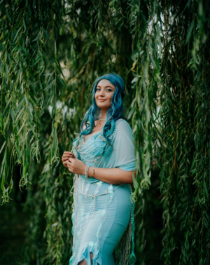 Frau mit blauen Haaren und funkelndem Kostüm steht unter einem Weidenbaum beim Elbenwald Festival. Die Mischung aus Musik und Fantasie zieht zahlreiche Besucher an, die sich verkleiden und die kreative Atmosphäre genießen.