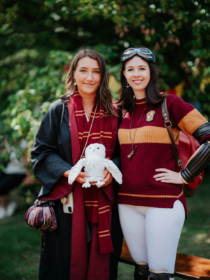 Zwei Frauen in Harry-Potter-Kostümen stehen lächelnd zusammen auf dem Elbenwald Festival. Die erste trägt einen Zaubererumhang und hält eine weiße Eule, während die zweite ein gestreiftes Sweatshirt mit Wappen trägt. Hintergrund mit grünen Bäumen.