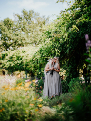 Zwei Frauen in fantasievollen Kostümen mit langen, hellen Haaren stehen in einer blühenden Gartenlandschaft auf dem Elbenwald Festival, umgeben von üppigem Grün und bunten Blumen, und strahlen Freude und Verbundenheit aus.