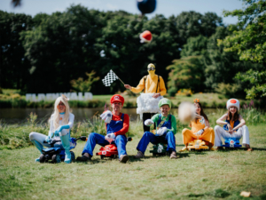 Sechs Cosplayer in bunten Kostümen sitzen auf kleinen Fahrzeugen am Elbenwald Festival. Die Gruppe zeigt Charaktere aus Videospielen, darunter Mario, Luigi und Toad, während ein Teilnehmer eine Zielflagge schwenkt. Fröhliche Atmosphäre am Ufer eines Teichs.