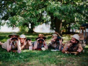 Fünf Personen in abenteuerlicher Kleidung liegen im Gras und beobachten mit Ferngläsern die Umgebung. Umgeben von Bäumen und einer entspannten Atmosphäre, verkörpern sie den kreativen Geist des Elbenwald Festivals.