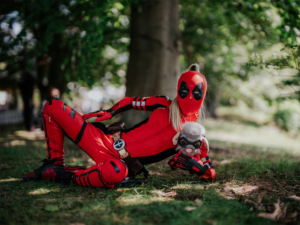 Ein Teilnehmer des Elbenwald Festivals in einem detaillierten Deadpool-Kostüm, der entspannt auf dem Gras liegt und eine Plüschfigur hält. Die Szene zeigt die kreative Atmosphäre des Festivals, das Musik und Nerdkultur vereint.