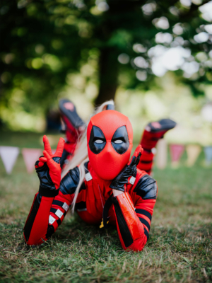 Cosplayer in einem roten Deadpool-Kostüm, der in einer entspannten Pose auf dem Gras liegt und das Peace-Zeichen zeigt. Im Hintergrund sind bunte Wimpel und verschwommene Bäume zu sehen, die die festliche Atmosphäre des Elbenwald Festivals unterstreichen.
