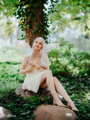 Frau mit langen, blonden Haaren und Schmetterlingsflügeln sitzt auf einem Stein im Grünen. Sie trägt ein elegantes, helles Kleid und wirkt verträumt. Die Szenerie ist von üppigem Blattwerk umgeben und strahlt eine magische Atmosphäre aus, passend zum Elbenwald Festival.