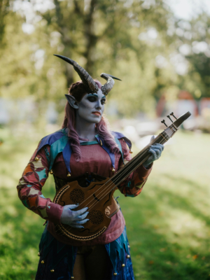 Kostümierte Person mit blauer Haut und Hörnern, die ein Musikinstrument hält, steht in einer grünen Umgebung beim Elbenwald Festival. Die lebendige Kleidung und die detailreiche Schminke unterstreichen den fantasievollen Charakter des Events.