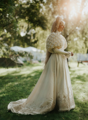 Eine elegant gekleidete Person in einem aufwendigen, weißen Kleid mit Spitze und Perlen, steht im Grünen während des Elbenwald Festivals. Die Sonne strahlt durch die Bäume und schafft eine magische Atmosphäre, die die fantasievolle Stimmung des Festivals unterstreicht.
