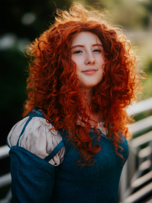 Eine junge Frau mit lockigem, rotem Haar trägt ein historisches Kostüm und lächelt in die Kamera. Der Hintergrund zeigt eine grüne Umgebung, die zur lebhaften Atmosphäre des Elbenwald Festivals passt. Perfekt für Fans von Cosplay und Musikfestivals.