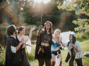 Eine Gruppe von Cosplayern in fantasievollen Kostümen feiert beim Elbenwald Festival. Die Person in der Mitte hält ein Schwert hoch, während die anderen applaudieren und jubeln, umgeben von einer malerischen Naturkulisse.
