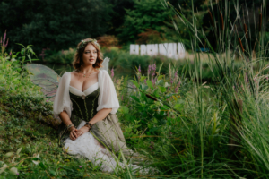 Eine Frau mit Elfenflügeln sitzt inmitten von bunten Blumen und grünem Gras beim Elbenwald Festival. Sie trägt ein elegantes, grünes Kleid und eine Blumenkrone, während im Hintergrund das Festival-Logo sichtbar ist.