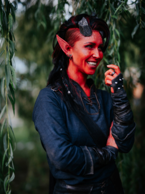 Eine lächelnde Person mit roten Gesichtstönen und spitzen Ohren, gekleidet in einem dunkelblauen Oberteil und schwarzen Handschuhen, steht unter einem Baum beim Elbenwald Festival. Die Atmosphäre ist festlich und kreativ, umgeben von grüner Natur.