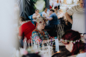 Basteln mit Damana Eine ältere Frau mit Blumenkranz und buntem Outfit bastelt an einem Tisch im Elbenwald Festival. Im Hintergrund sind weitere Teilnehmerinnen und Teilnehmer mit verschiedenen Bastelprojekten zu sehen, die die kreative Atmosphäre des Festivals widerspiegeln.
