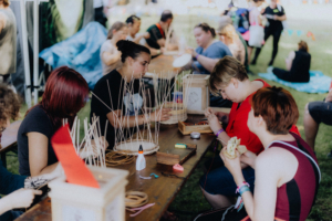 Basteln mit Damana Teilnehmende am Elbenwald Festival basteln an einem kreativen Workshop. Auf einem Tisch liegen Materialien wie Kleber, Schnüre und Holzrahmen. Die Atmosphäre ist entspannt und gemeinschaftlich, während die Teilnehmer ihre kreativen Projekte umsetzen.