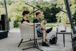 darkviktory & Kostas Kind Zwei junge Männer sitzen auf einer Bühne beim Elbenwald Festival und diskutieren. Der eine hält ein Mikrofon, während der andere aufmerksam zuhört. Im Hintergrund sind grüne Bäume und Technik sichtbar, die die Festivalatmosphäre unterstreichen.