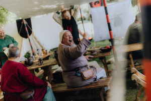Die Roten Adler Eine fröhliche Gruppe von Teilnehmern beim Elbenwald Festival, die in historischer Kleidung an Holztischen sitzt, während sie lachen und essen. Im Hintergrund sind dekorative Elemente und weitere Festivalbesucher zu sehen, die die lebendige Atmosphäre des Events unterstreichen.
