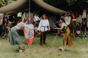 Die roten Adler Zwei Frauen und ein Mann interagieren auf dem Elbenwald Festival, während sie an einem Seil ziehen. Im Hintergrund sind Zelte und bunte Stände zu sehen, die die festliche Atmosphäre eines nerdigen Musikfestivals und einer Convention widerspiegeln.