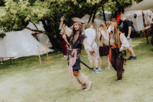 Ein Pirat in Kostüm mit Flasche in der Hand, umgeben von Festivalbesuchern in bunten Outfits, beim Elbenwald Festival. Im Hintergrund Zelte und eine entspannte Festivalatmosphäre auf einer grünen Wiese.