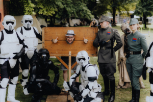 501stLegion Eine Gruppe von Cosplayern in Star Wars-Kostümen posiert auf dem Elbenwald Festival. Im Vordergrund steht ein Mann in einem Holzgestell, der Daumen hoch zeigt, während die Charaktere um ihn herum stehen und die Festivalatmosphäre genießen.