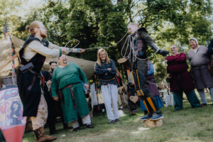 Die Roten Adler vs. 501st Zwei Teilnehmer des Elbenwald Festivals führen eine humorvolle Vorführung auf, während eine Gruppe von Zuschauern in mittelalterlicher Kleidung zuschaut. Die Szene zeigt interaktive Unterhaltung und die lebendige Atmosphäre des Festivals.