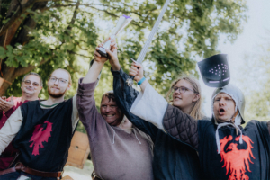 Die Roten Adler vs. 501st Gruppierung von Festivalbesuchern in mittelalterlicher Kleidung, die jubelnd Pokale und Schwerter in die Höhe halten. Bäume im Hintergrund schaffen eine festliche Atmosphäre beim Elbenwald Festival, das Musik und Nerd-Kultur verbindet.