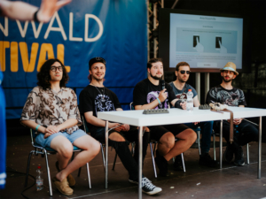 The Nerds Paneldiskussion auf dem Elbenwald Festival mit fünf Sprechern, die auf einer Bühne sitzen. Im Hintergrund ist ein Bildschirm mit Anweisungen zu sehen. Die Atmosphäre ist entspannt und kreativ, ideal für Fans von Musik und Nerdkultur.