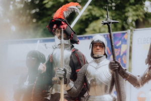 Zwei Ritter in historischer Rüstung und ein verkleideter Charakter stehen auf dem Elbenwald Festival. Im Hintergrund sind bunte Banner zu sehen, die das kreative und fantasievolle Ambiente des Festivals widerspiegeln.