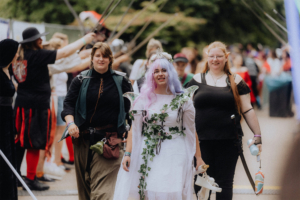 Drei Frauen in Kostümen beim Elbenwald Festival, eine von ihnen trägt ein feenhaftes Outfit mit lila Haaren und Blumenkranz. Der Hintergrund zeigt eine lebhafte Festivalatmosphäre mit Teilnehmern in verschiedenen Fantasiekostümen und Requisiten.