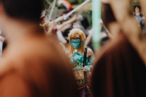 Ein Cosplayer in einem bunten Outfit, inspiriert von einem Fantasy-Charakter, steht im Mittelpunkt des Elbenwald Festivals. Die Veranstaltung zieht zahlreiche Fans von Musik und Popkultur an, während im Hintergrund andere kostümierte Teilnehmer zu sehen sind.