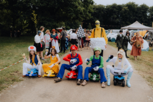 Gruppenkostümierte Teilnehmer beim Elbenwald Festival, die in kleinen Karts sitzen. Sie verkörpern Charaktere aus Videospielen, darunter Mario, Luigi und Prinzessin Peach, umgeben von Festivalbesuchern und Zelten im Hintergrund.